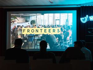 A dark room with a Fronteers screen in the front, due to the light contrast of the screen in the dark room there black silhouets visible of the people sitting in front of the screen. The room is also covered in a blue glow of a neon light that says the word 'whale' and has a small stylized whale icon next to it